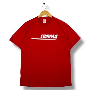 Peut inclure: T-shirt rouge avec le mot "COMPAQ" en blanc et une ligne blanche horizontale en dessous. Le t-shirt est suspendu à un cintre en bois sur fond blanc. Col rond et manches courtes.