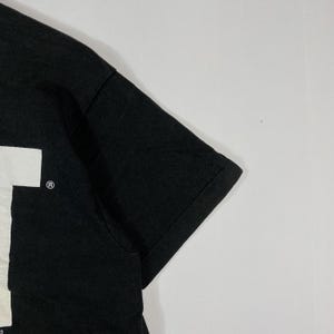 May include: Black short-sleeved t-shirt with a white graphic on the front. The shirt has a crew neck and a classic fit. The sleeve is visible in the image.