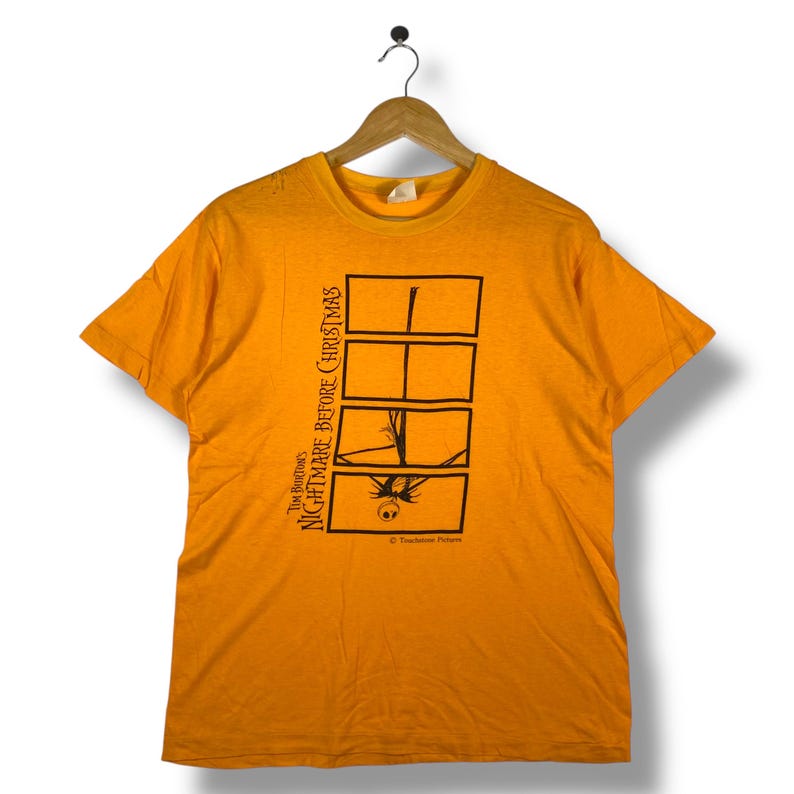Puede incluir: Camiseta naranja con un dise&ntilde;o gr&aacute;fico de "Pesadilla antes de Navidad" de Tim Burton. El dise&ntilde;o incluye paneles rectangulares negros con im&aacute;genes de Jack Skellington. El texto "Tim Burton's Nightmare Before Christmas" est&aacute; impreso verticalmente.