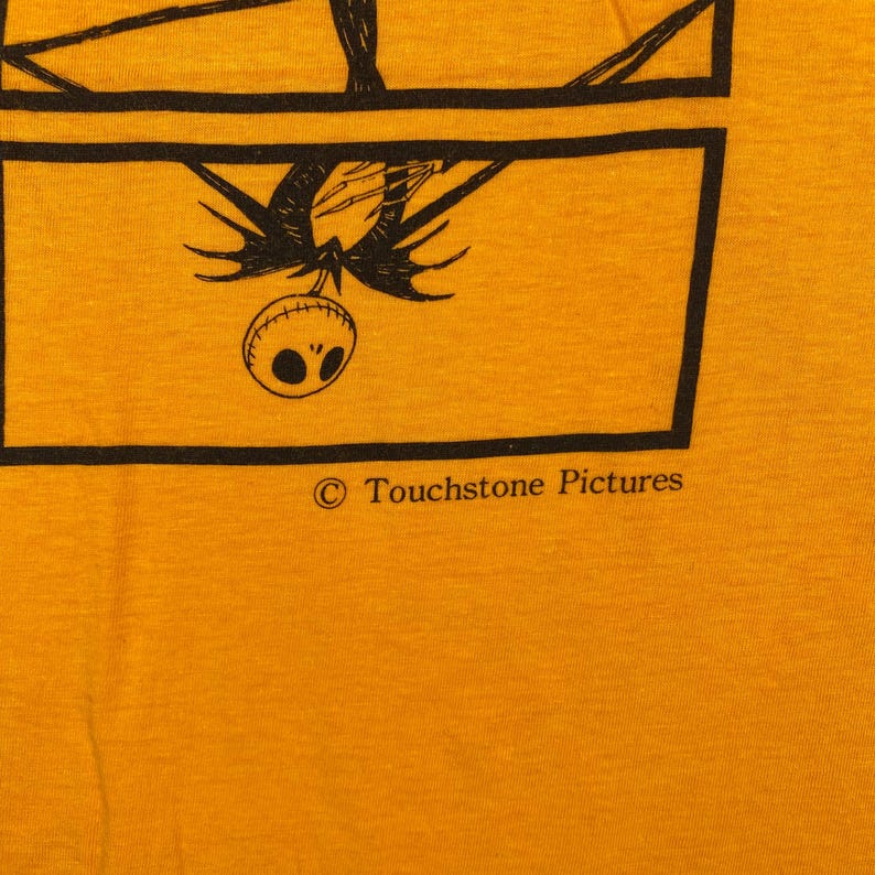 Puede incluir: Camiseta amarilla mostaza con un gr&aacute;fico negro de un personaje de Pesadilla antes de Navidad. El gr&aacute;fico est&aacute; enmarcado por l&iacute;neas negras. El texto "&copy; Touchstone Pictures" est&aacute; impreso debajo.