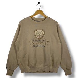 Könnte beinhalten: Beige Sweatshirt mit gesticktem Text "CALIFORNIA STATE UNIVERSITY San Bernardino". Das Design beinhaltet ein Wappen. Das Sweatshirt hat einen Rundhalsausschnitt und lange Ärmel.