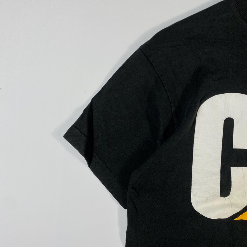 May include: Black t-shirt with a large white letter "C" and yellow accents. The short-sleeved shirt is made of a dark fabric and is partially visible in the image. The shirt's design is bold and eye-catching.