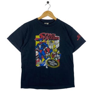 Puede incluir: Una camiseta negra con un diseño de estilo cómic. El gráfico incluye las palabras "SPACE FUTURE" en rojo y blanco, junto con ilustraciones de personajes disfrazados. La camiseta también tiene el texto "60st ADVENTURE STORY".
