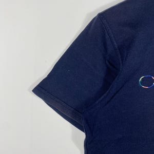 May include: Navy blue short-sleeved t-shirt with a small, multicolored embroidered design on the chest. The shirt has a classic crew neck and a simple, clean design, suitable for casual wear.