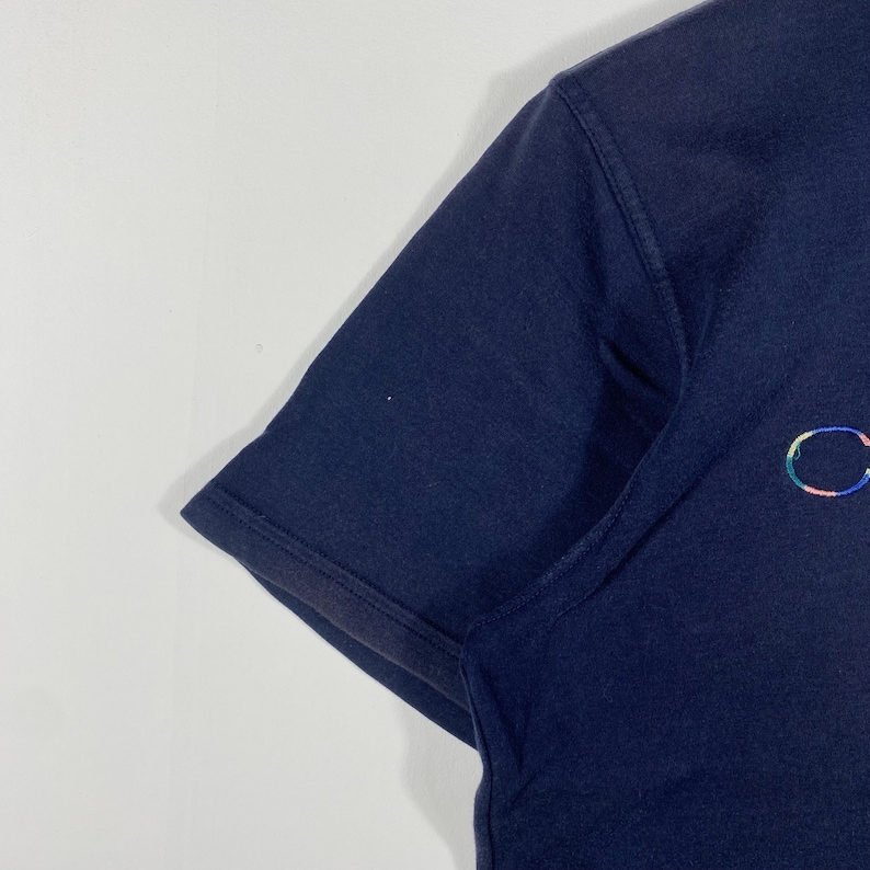 May include: Navy blue short-sleeved t-shirt with a small, multicolored embroidered design on the chest. The shirt has a classic crew neck and a simple, clean design, suitable for casual wear.