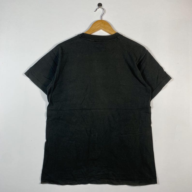May include: A black t-shirt hanging on a wooden hanger. The short-sleeved shirt is made of a soft material and has a slightly faded appearance. The shirt is displayed against a plain white background.