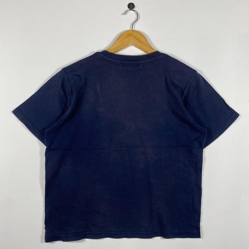 May include: A navy blue short-sleeved t-shirt hanging on a wooden hanger. The shirt is made of a soft material and has a simple design. The shirt is displayed against a white background.