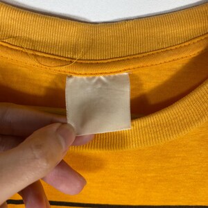 Puede incluir: Primer plano de un cuello de camiseta amarillo mostaza con una etiqueta blanca en blanco. La etiqueta es rectangular y parece estar hecha de tela. El cuello de la camisa es acanalado y la tela tiene un aspecto ligeramente desgastado.