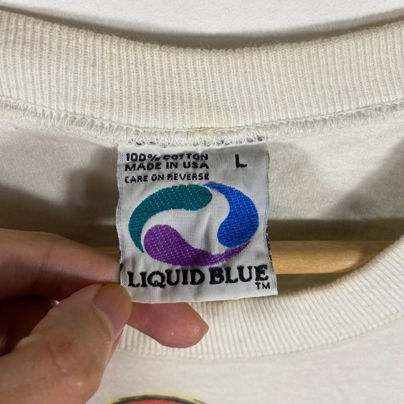 May include: Close-up of a white t-shirt tag with the text "100% COTTON MADE IN USA CARE ON REVERSE" and the Liquid Blue logo. The tag also has a size "L" label.
