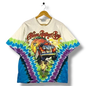 May include: White tie-dye t-shirt featuring a graphic of a vintage car, a peach, and the text "Allman Brothers Band." The shirt has a colorful tie-dye pattern on the sleeves in shades of blue, green, and purple.