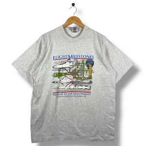 May include: Light gray t-shirt featuring a graphic of various aircraft with the text "FLIGHT MILESTONES" and "National Air and Space Museum Smithsonian Institution, Washington, DC". The shirt has a crew neck and short sleeves.