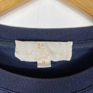 May include: Close-up of a navy blue t-shirt with a tag. The tag is off-white with the text "courr&egrave;ges 2 paris" and the size "S". The shirt is made of a soft material.