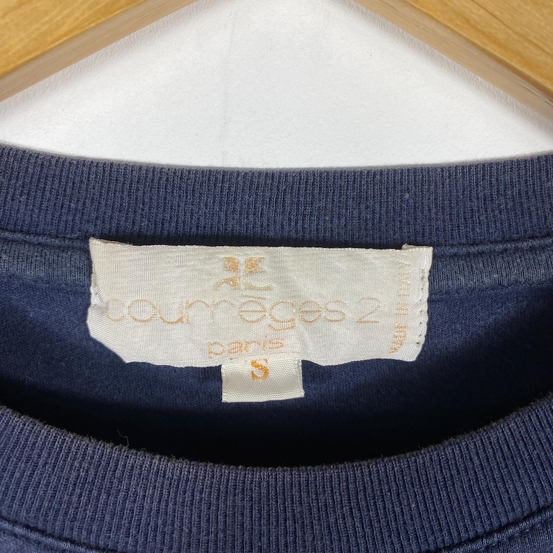 May include: Close-up of a navy blue t-shirt with a tag. The tag is off-white with the text "courr&egrave;ges 2 paris" and the size "S". The shirt is made of a soft material.