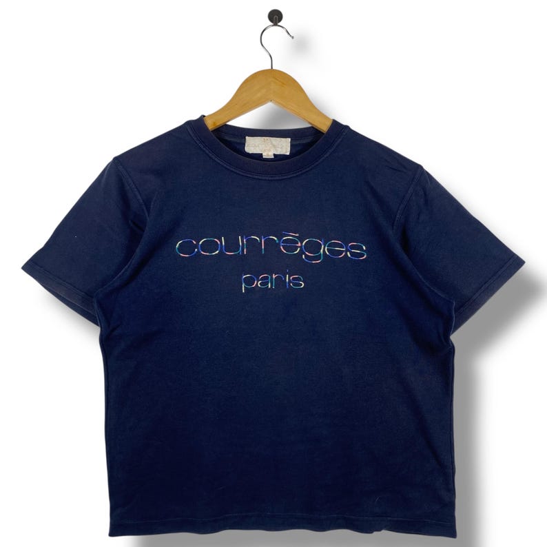 May include: Navy blue t-shirt with the text "courr&egrave;ges paris" in iridescent thread. The shirt has short sleeves and a crew neck. The shirt is hanging on a wooden hanger.