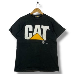 May include: Black t-shirt with the white "CAT" logo and a yellow triangle. The shirt is hanging on a wooden hanger. The Caterpillar logo is printed on the front of the shirt.