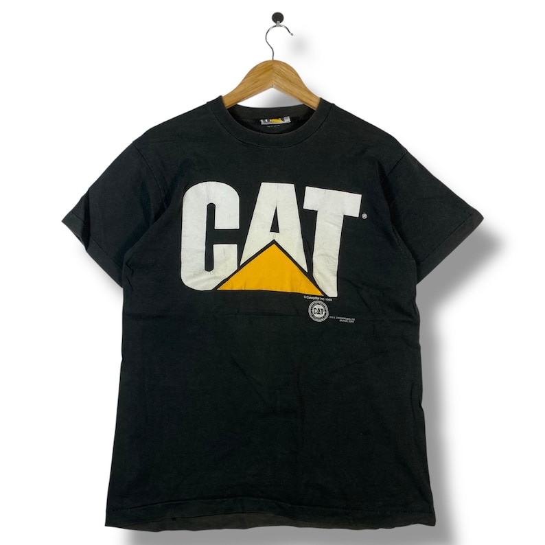 May include: Black t-shirt with the white "CAT" logo and a yellow triangle. The shirt is hanging on a wooden hanger. The Caterpillar logo is printed on the front of the shirt.