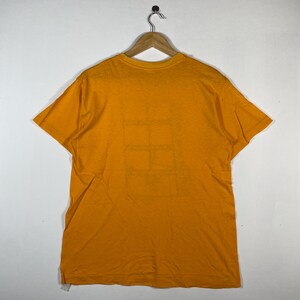 Puede incluir: Una camiseta de color amarillo mostaza colgada de una percha de madera. La camiseta tiene un dise&ntilde;o geom&eacute;trico simple en la parte delantera. La tela parece ser un material suave y ligero, adecuado para ropa informal.