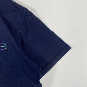 May include: Navy blue short-sleeve t-shirt with a small, iridescent logo on the chest. The shirt's sleeve is visible, showing the hem and the texture of the fabric. The background is plain white.