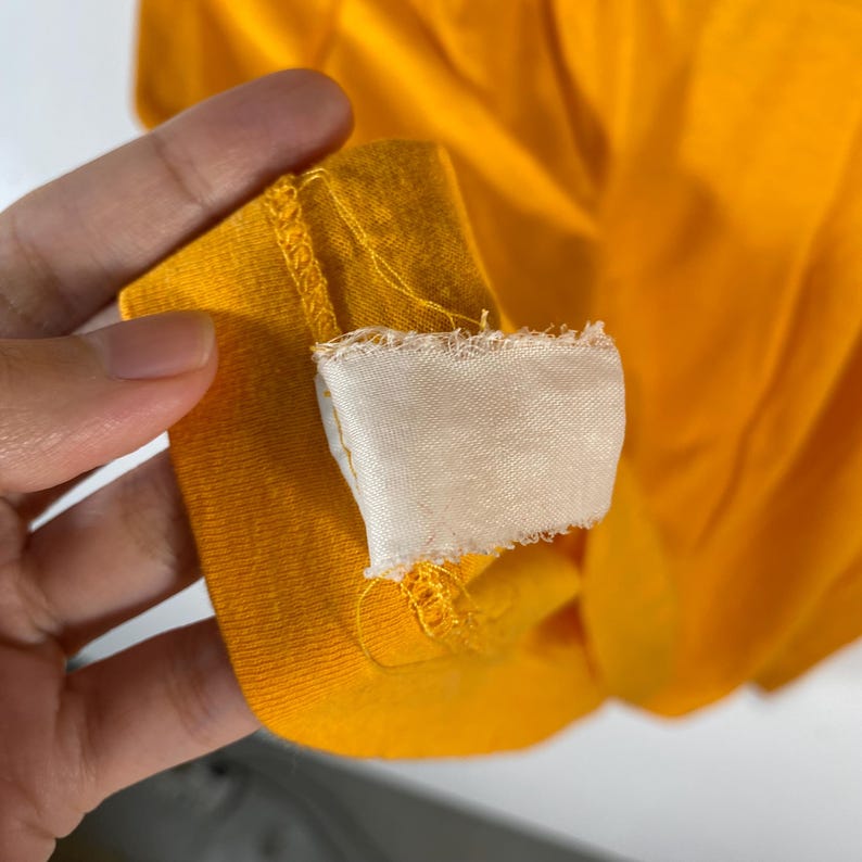 Puede incluir: Primer plano de una camiseta amarilla brillante con una etiqueta blanca adjunta. La etiqueta tiene un borde &aacute;spero e inacabado. La tela es lisa y parece ser un material ligero. La camiseta es parcialmente visible, con el foco en la etiqueta y la tela.