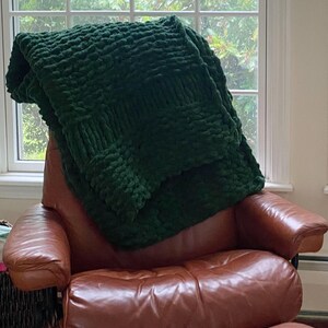 May include: A dark green, chunky knit blanket draped over a brown leather armchair with a matching footstool. The blanket has a textured, woven appearance. The chair is positioned in front of a window with a view of green foliage.