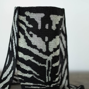 May include: Black and silver beaded shoulder bag with a zebra stripe pattern. The bag has a cylindrical shape with a flat bottom and a long strap. The bag is made of small, shiny beads.