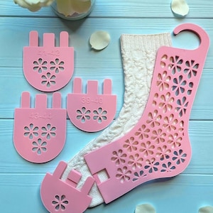 May include: A collection of pink sock shapers with floral cutouts and size markings, next to a cream cable-knit sock. The shapers are marked with sizes 41-42, 39-40, and 43-44. The background is a light blue surface with white flower petals and a vase of white roses.