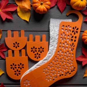 Puede incluir: Un molde de calcetín naranja con diseño floral y un calcetín gris de punto trenzado, rodeado de calabazas y hojas de otoño. El molde de calcetín tiene indicadores de talla: 37-38, 41-42, 43-44 y 39-40.