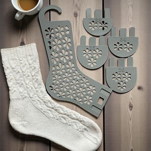 Puede incluir: Un calcetín blanco de punto trenzado, una forma de calcetín gris con recortes florales y cuatro medidores de talla de calcetín grises (33, 34, 35, 36) están dispuestos sobre una superficie de madera. Una taza de café está en la esquina superior izquierda.