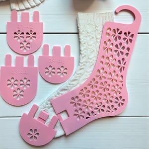 Puede incluir: Hormas de calcetines rosas con cortes florales e indicadores de talla, junto a un calcetín blanco de punto trenzado. Las hormas muestran tallas como 41-42, 39-40 y 43-44. También hay una taza de café y un suéter blanco.