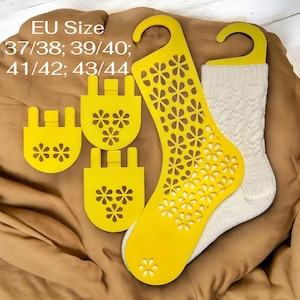 Op de afbeelding: Crèmekleurige kabelgebreide sokken met gele sokkenhangers en maattabellen. De gele hangers hebben een bloemvormig uitgesneden ontwerp. De tekst "EU Size 37/38; 39/40; 41/42; 43/44" is zichtbaar.