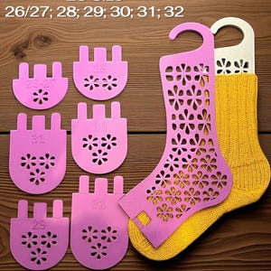 May include: A display of pink sock shapers with flower cutouts, alongside a yellow sock and a white sock shaper. The text "EU Size 26/27; 28; 29; 30; 31; 32" is visible. The items are arranged on a wooden surface.