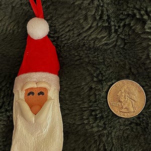 Könnte beinhalten: Ein Weihnachtsschmuck in Form eines Weihnachtsmanns mit roter Mütze und weißem Bart. Der Schmuck hat eine Schlaufe zum Aufhängen und liegt neben einer US-Vierteldollarmünze.
