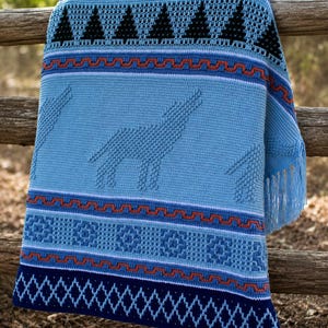May include: A light blue crocheted blanket with a wolf design and geometric patterns. The blanket features a fringe and is draped over a wooden fence. The design includes black triangles, orange and blue zigzags, and diamond patterns.