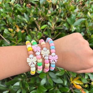 May include: Four beaded bracelets in pastel colors, each adorned with a bow. The bracelets feature yellow, pink, green, and blue beads. The bows are white and pink, adding a decorative touch to the jewelry.