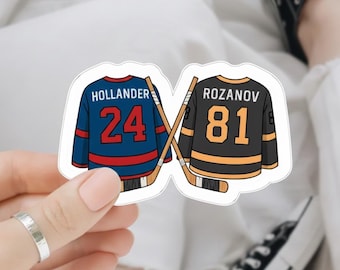 May include: A sticker featuring two hockey jerseys with crossed hockey sticks. One jersey is blue with "HOLLANDER 24", the other is black with "ROZANOV 81". The sticker is held by a person.