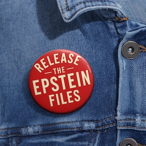 May include: A red button with white text that reads "RELEASE THE EPSTEIN FILES" pinned to a denim jacket. The button is round and has a glossy finish. The jacket is a medium wash denim.