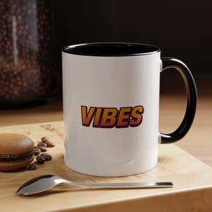 Può includere: Tazza in ceramica bianca con interno e manico neri. La parola "VIBES" è stampata in lettere sfumate arancioni e gialle. Un cucchiaio, un macaron e chicchi di caffè sono su una superficie di legno.