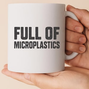 May include: White ceramic mug with a curved handle. The mug features the text "FULL OF MICROPLASTICS" in a bold, black, sans-serif font. The mug is held by a person.