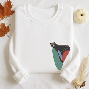 Puede incluir: Sudadera blanca con un gráfico de un oso negro descansando sobre una forma abstracta colorida. El oso es marrón y la forma abstracta es verde, azul y rojo. La sudadera tiene mangas largas y cuello redondo.