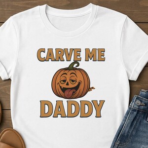 May include: White t-shirt featuring a smiling orange pumpkin with a green stem and a protruding tongue. The text "CARVE ME" is above the pumpkin, and "DADDY" is below, all in a brown, outlined font. The shirt is paired with blue jeans.