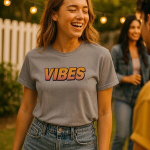 May include: A woman wearing a grey t-shirt with the word "VIBES" in orange and red lettering. She is wearing denim shorts. The background includes a white fence, string lights, and other people.
