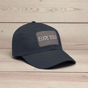Puede incluir: Gorra de béisbol azul marino oscuro con visera curva. Un parche rectangular gris en la parte delantera muestra el texto blanco "FUCK YOU". La gorra está sobre un fondo de vetas de madera clara.