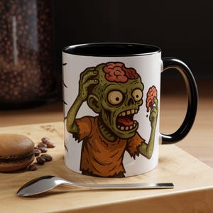 May include: White ceramic mug with a black interior and handle, featuring a cartoon zombie illustration. The zombie is green with exposed brains and wearing an orange shirt. A spoon and a pastry are in the foreground.