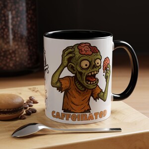 May include: White ceramic mug with a black interior and handle. The mug features a cartoon zombie illustration with the word "CAFFEINATED" in orange. The zombie is green with exposed brain matter and wearing an orange shirt.