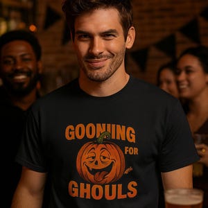 May include: Black t-shirt with the text "GOONING FOR GHOULS" and a smiling jack-o'-lantern graphic in orange. The shirt is worn by a person in a bar setting, with other people and a beer glass in the background.