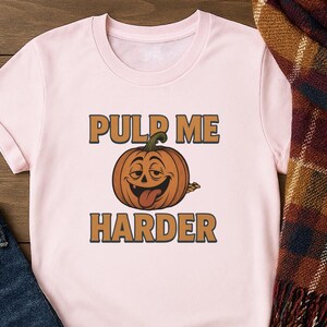 May include: A light pink t-shirt with the text "PULP ME HARDER" in brown letters. A cartoon pumpkin with a smiling face and tongue sticking out is in the center of the text.