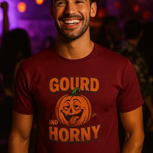 May include: Maroon t-shirt with the words "GOURD AND HORNY" in orange, along with a cartoon pumpkin graphic. The pumpkin has a green stem and a playful expression. The shirt is worn by a smiling person.