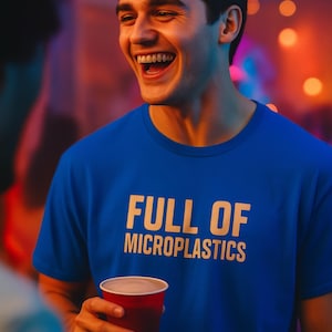 May include: A blue t-shirt with the text "FULL OF MICROPLASTICS" in white. The person is holding a red cup and smiling. The background is blurred with warm colors.