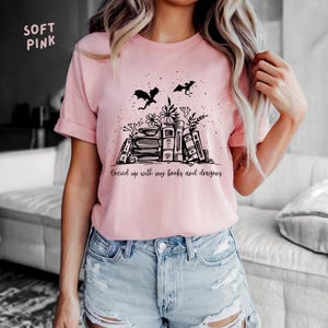 May include: Pale pink t-shirt featuring a black illustration of books, flowers, and dragons. The text "Cozied up with my books and dragons" is printed below the graphic. The shirt is made of a soft material.