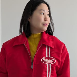 May include: A red jacket featuring a white stripe and a "Peterbilt Toronto" logo. The jacket has a zip closure and a collar. A yellow turtleneck is worn underneath. The jacket is a vibrant piece of outerwear.